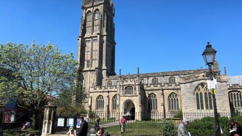 Image of St John the Baptist's Church on a busy high street. There is a blue sky behind the church and in the foreground there is greenery, an iron gate and some people. 