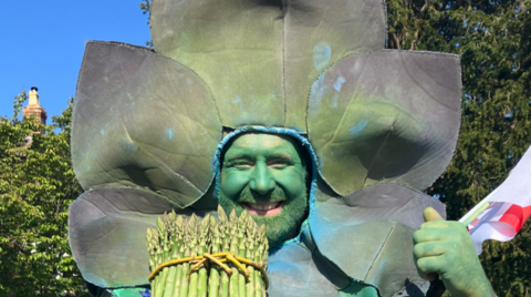 A man with a painted green face is wearing a green asparagus costume and holding asparagus in his right hand. He is smiling and trees are behind him.