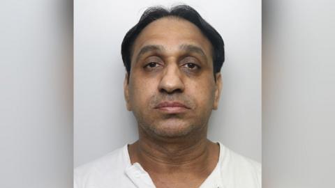 A mugshot of Bostan, an Asian male with dark wavy hair and brown eyes. He is wearing a white shirt and has some light facial stubble.