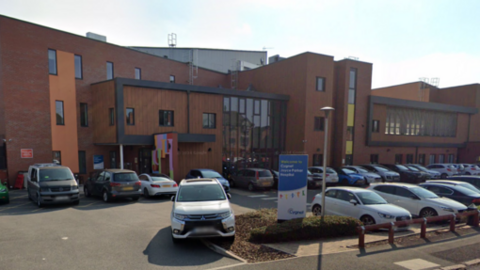 The outside of a hospital, with a car park in front of it. The hospital building is red brick with black frames around the centre structure. 