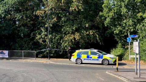 Police car and tape across a wooded area off a roundabout with a sign to Swaythling.