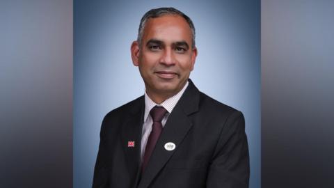 Adi Malviya is wearing a dark-coloured suit with a striped shirt and burgundy red tie. He is smiling and looking at the camera.