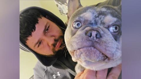 Peter Macann a man with short black hair. He is looking at the camera and is wearing a grey hoodie with the hood up. Next to him is a grey speckled French bulldog who is also looking at the camera. 