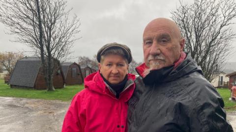 A woman wearing a flat cap and a pink/red hooded coat and a bald man in a black hooded coat. They are standing outdoors in a caravan park, with a tree and row of cabins behind them.
