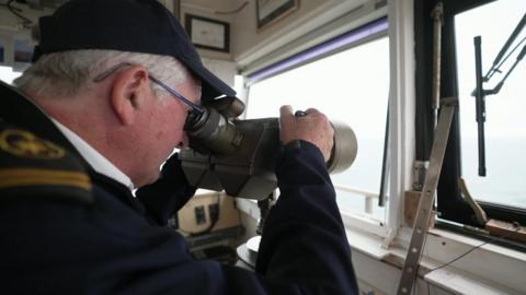 Jerry wears a dark cap and top with gold epaulettes. He is looking through a telescope through a window out to sea.