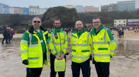 Llun o 4 gwirfoddolwr gyda elusen St John's Ambulance Cymru yn sefyll yn y mor ar draeth Dinbych y Pysgod. Mae'r 4 yn gwisgo siacedi llachar melyn. Mae llawer o bobl yn y cefndir ar y traeth.