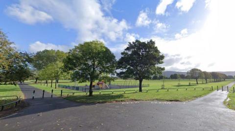 A wide view of a green space in Eston Recreation Ground. Grassy areas are intersected with grey pavement. A small children's play park is seen in behind two trees.