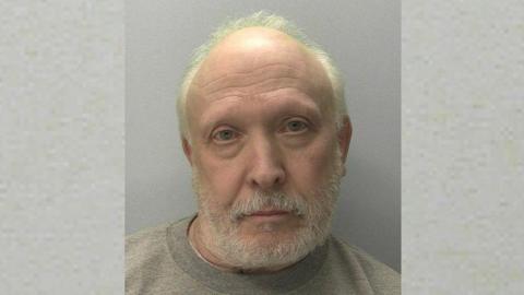 Police mugshot of Richard Law. He has white, receding hair and a white beard and he is wearing a grey jumper.