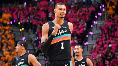 Victor Wembanyama, wearing a black San Antonio vest with a turquoise, pink, and orange stripe across the chest, punches his right fist in celebration and smiles