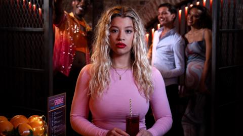 A young woman with bleached blonde shoulder length hair wears a pink top and holds a drink in a glass with straw held in two hands at waist level. Behind her a group of friends takes a selfie in a dimly lit bar or nightclub.ford, who plays Anna Knight