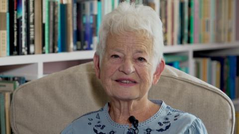Jacqueline Wilson looks at the camera, sat on an armchair. She is wearing a blue blouse and is sat infront of a bookcase
