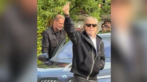 Paul McCartney waves to the crowd. He is smiling and wearing sunglasses. He is wearing a light coloured top, black jeans and a black hooded jacket. He is standing in front of a car and some people can be seen in the background, including someone filming on their mobile phone.