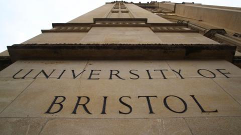 A shot pointing up the height of a University of Bristol building. 'The University of Bristol' can be seen spelled out in the beige stone of the building's wall which reaches up to the sky.