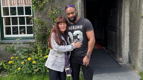 Denise, with red hair and a light blue jacket, with her arms around her partner Sylvester Abayomi, wearing a black T-Shirt with white numbers and with sunglasses on his head, in a photo released by the family.