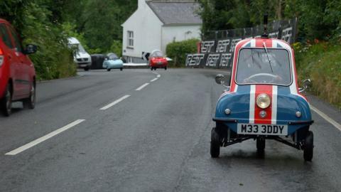 The small Peel P50, with a painted red, white and blue union jack flag drives up hill on a wet road. In the distance, two Tridents, one pale blue, one red follow behind. There's TT related signage next to the road.