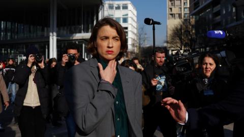 Adèle Haenel wearing a dark grey suit jacket and a dark green shirt underneath. She is standing outdoors, squinting in the sunshine, and is surrounded by journalists taking photos of her