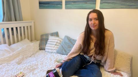 Lara is sitting on her bed with her legs tucked underneath her. She has long brown hair. She is wearing a pink cardigan and blue jeans. On the bed is a packet of antidepressants and a cardboard box.