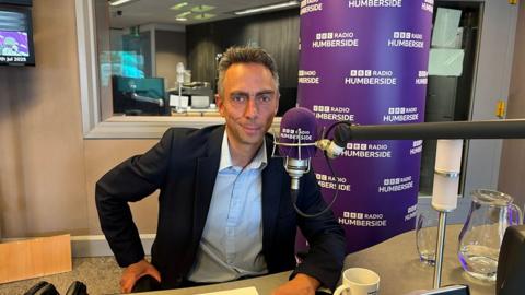 Mike Ross with short dark hair a dark jacket and blue shirt sits behind a microphone in the BBC Radio Humberside studio