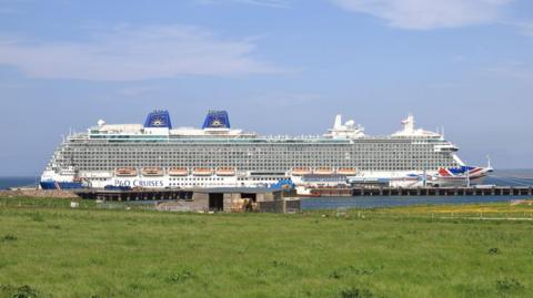 A large P&O cruise ship, Britannia, moored in Kirkwall in June 2023. A field of grass can be seen in the foreground.
