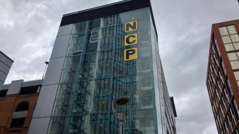 A tall glass building which has a sign that says NCP in black lettering against a yellow background. Behind the building is a cloudy, grey sky