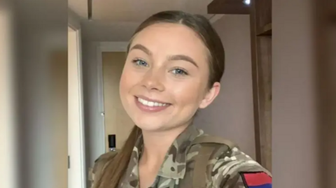 Jaysley Beck smiles at the camera in her camouflage army uniform. She has her brown hair tied back in a ponytail