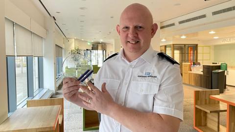 Mark Vipond holds his circular, glass award. He has a shaved head and is wearing a white NHS branded shirt with blue epaulettes