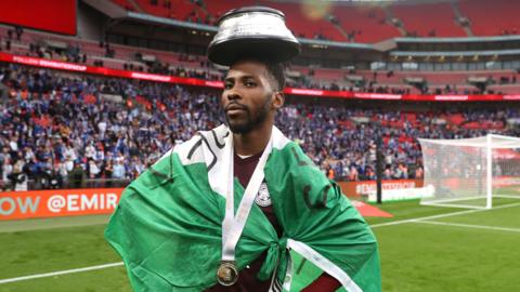 Leicester City forward Kelechi Iheanacho poses inside Wembley Stadium with the FA Cup trophy plinth on his head, a Nigeria flag draped around his shoulders and a medal around his neck