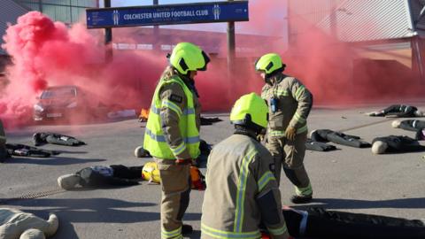 A simulated explosion in a car gives off red smoke while a group of firefighters attend to a large group of mannequins that are being used as 'casualties' in a terrorism training exercise by the London Fire Brigade.