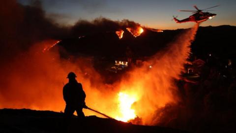 The silhouette of a firefighter is seen against bright orange flames as a helicopter drops water on burning hills in the distance 