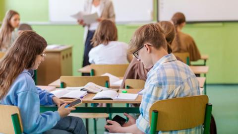 A stockshot of a teenage boy and girl in a classroom at a desk with textbooks laid out and they are looking at their mobile phones. The girl has long brown hair, wearing a blue jumber and jeans. The boy has golden brown hair and is wearing black glasses and a blue and white shirt. 