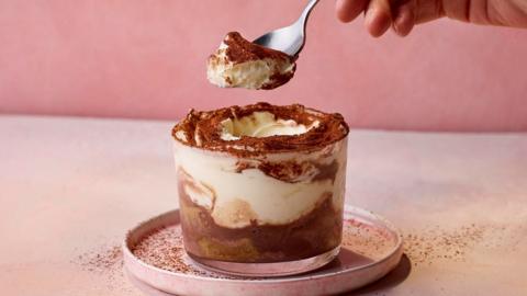 A person takes a spoonful out of a tiramasu in a small mug. It has chocolate and cream and a sprinkling of cocoa on the top.