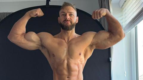 Brendan Pearl is 35 and has light brown hair across the top of his head, he has matching colour beard and moustache and is topless, and has very large muscles. 