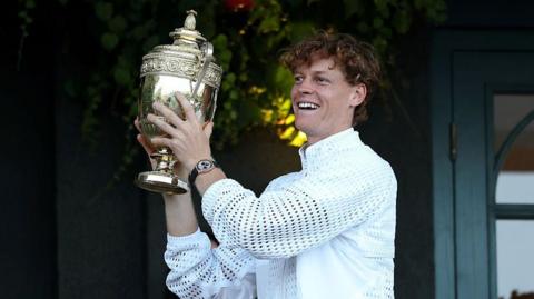 Jannik Sinner of Italy shows The Gentlemen's Singles Trophy to spectators from a balcony of Centre Court. He is smiling and holding the trophy up. He is wearing a white jacket and a watch with a gold face and black wrist strap. The trophy is gold.