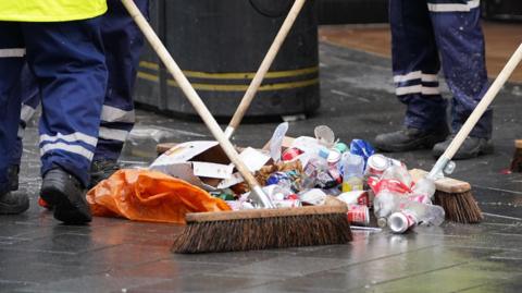 Four men with brooms sweeping cans and bottles