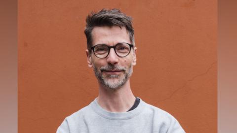 Tim Guy is standing in front of a bright orange wall. He is looking directly at the camera and smiling. He is wearing a pair of round glasses and a grey pullover.