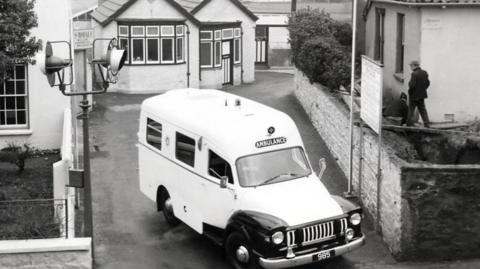 A black and white image of a very old fashioned white ambulance as it exits a driveway. There are houses on either side and a building with bay windows behind it.