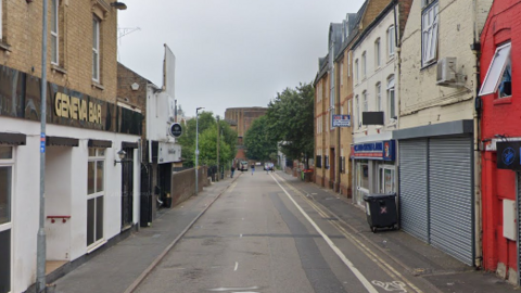 Geneva Street in Peterborough, showing Geneva Bar to the left, with a building to the right with metal shutters across its windows and a large black wheelie bin outside. A takeaway shop is next door. The single lane road is empty, with no traffic or people.  