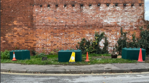Three green concrete blocks sit on a grass verge on the side of a road - there are traffic cones in front of them and a brick wall behind.