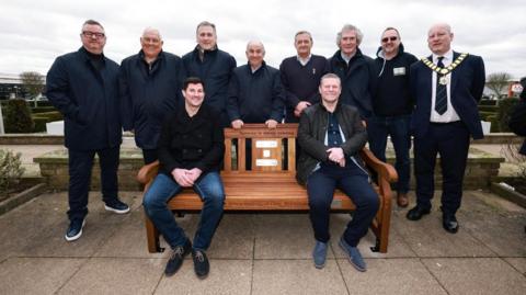 Several people gather around a brown wooden bench. Eight people are stood behind it, with two people sat on the bench. On the bench there are several plaques, and above them is some black writing which says "someone is always listening".