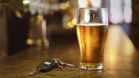 A pint of amber coloured beer sitting on a wooden bar with a set of keys next to it and blurred lights in the background