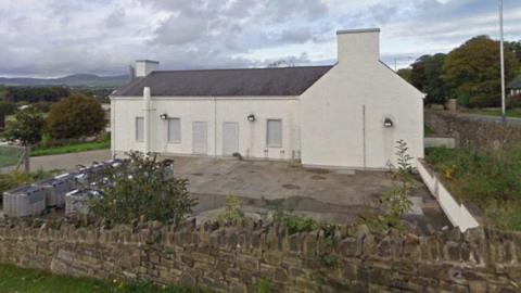 The pumping station which is a white building that resembles a large house with shutters windows and doors. It is surrounded by a Manx stone wall and there are industrial packs within its grounds. There is greenery surrounding the site.