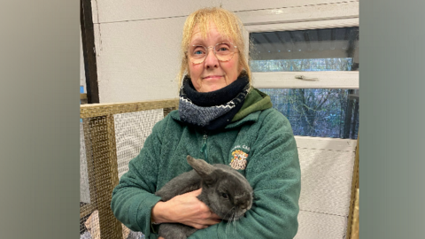 A woman stands smiling at the camera holding a grey rabbit 