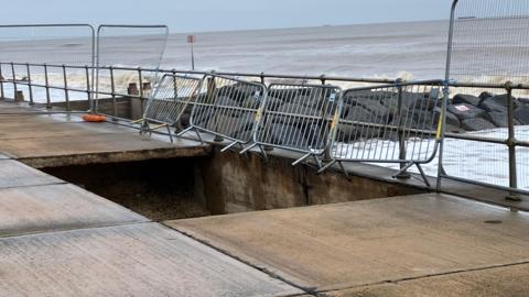 A large rectangular-shaped hole surrounded by metal fencing in the seafront walkway. A grey sky and muddy brown North Sea are in the background. 