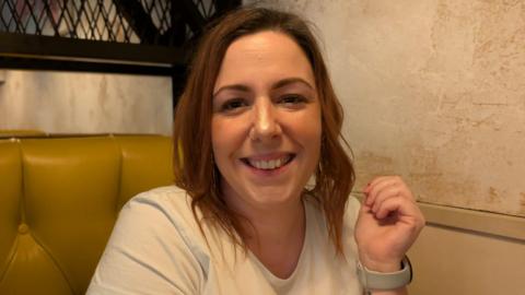 A woman with shoulder length red hair and a nose ring wearing a white t-shirt is smiling at the camera. She appears to be sitting in a booth at a restaurant.