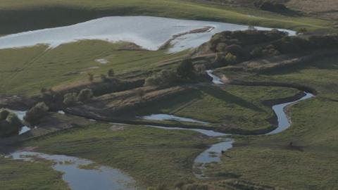 A river in a green fields, which swirls off course into more green fields in a bendy shape. 