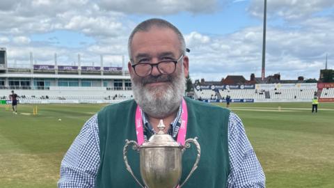 A man in a long sleeve shirt and green vest top poses with a trophy against the backdrop of a cricket ground. He has cropped hair and is wearing black glasses that sit on the end of his nose. 