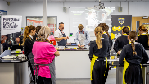 Teachers stand at the front of the classroom delivering a cooking class. They are wearing shirts and aprons. Pupils are facing towards the teachers. They have their back to the camera. Posters and a whiteboard are on display in the classroom.