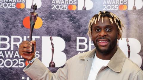 Myles Smith is standing on the right of the picture, holding his Brit Award. The award is the shape of a person, and is wooden. Myles has brown and blonde hair, a white tshirt, and beige jacke. He is standing in front of a backdrop which says BRIT AWARDS 2025 across it. 