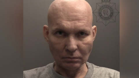 A man with a bald head is wearing a grey top and staring into a police custody picture.
