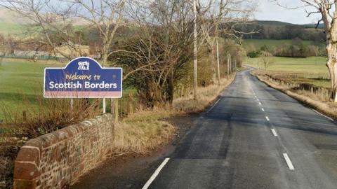 A sign saying Welcome to the Scottish Borders next to a long, straight road with trees and fields around it